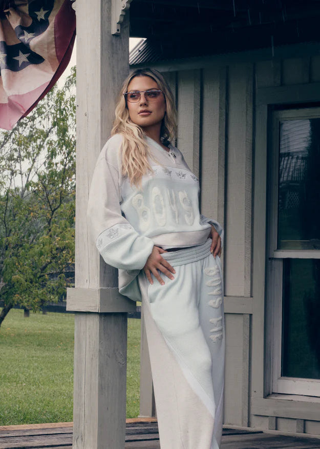 Woman in a light-colored outfit standing on a porch with an American flag in the background. Boys lie lounge set. private school set. blue. local business. boys lie. Fayetteville arkansas. university of arkansas.