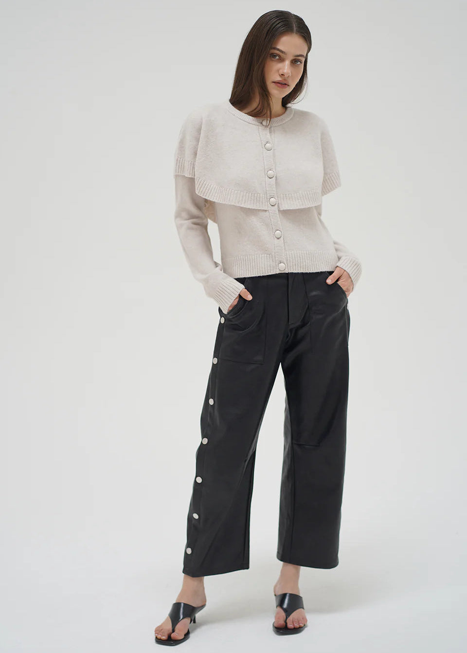 Woman wearing a beige cardigan and black pants with button details on a white background