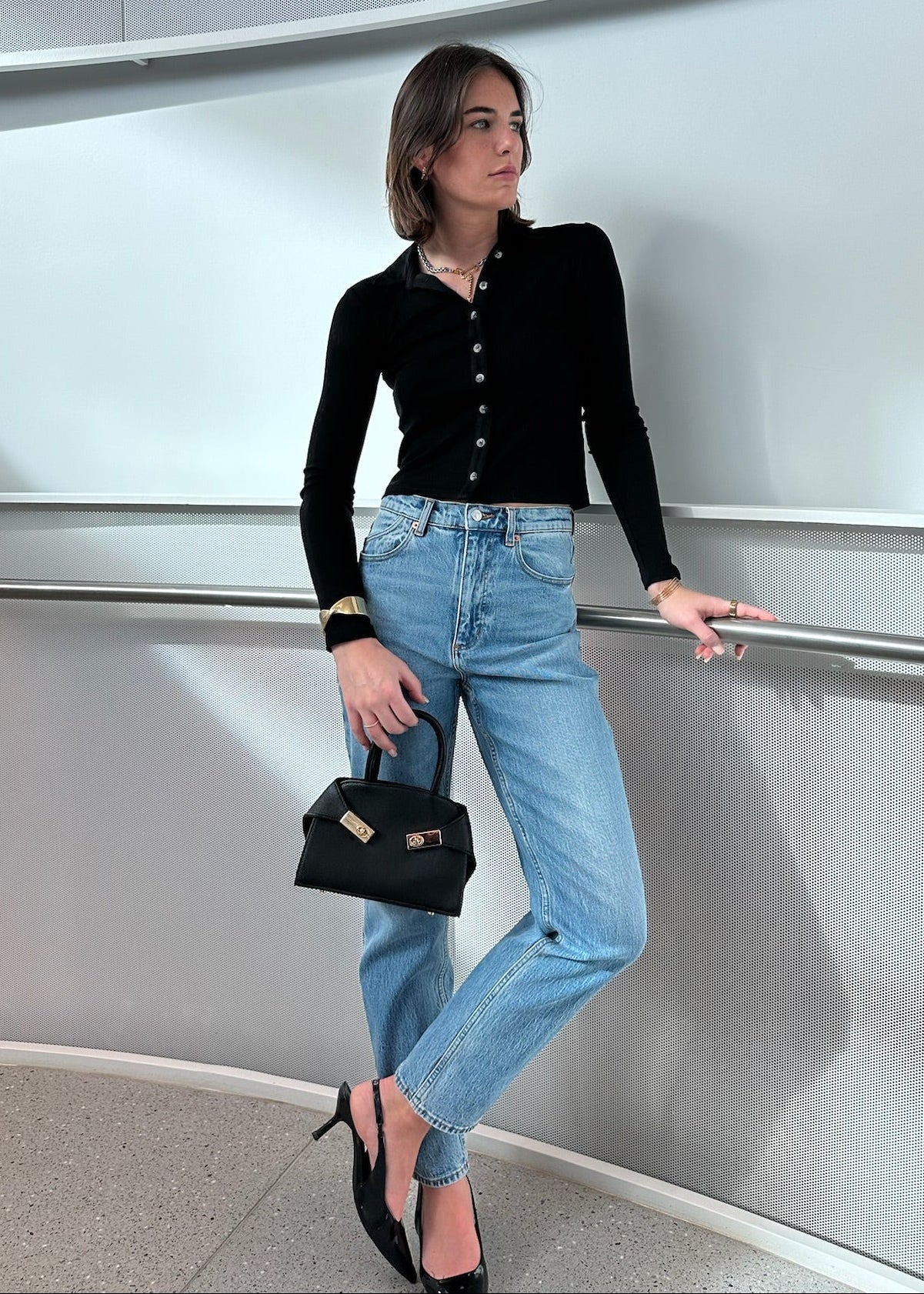 Woman in black top and blue jeans holding a black handbag, standing against a metallic wall.