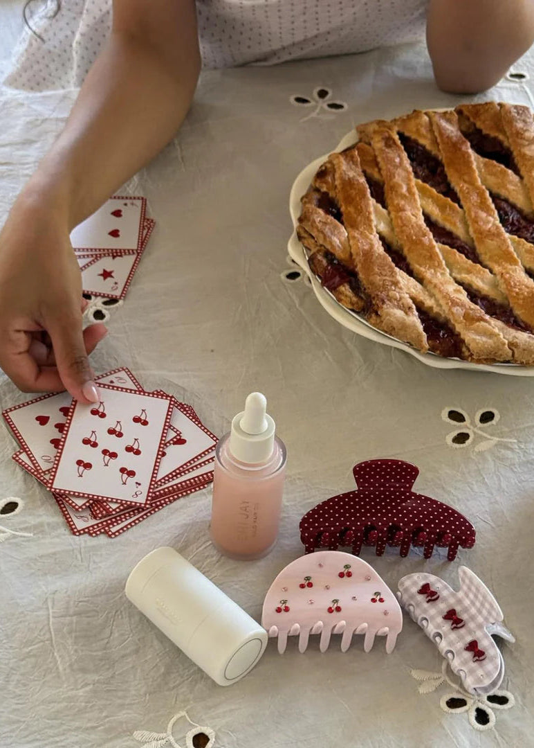 Person playing cards with a pie, hair accessories, and a bottle on a tablecloth.