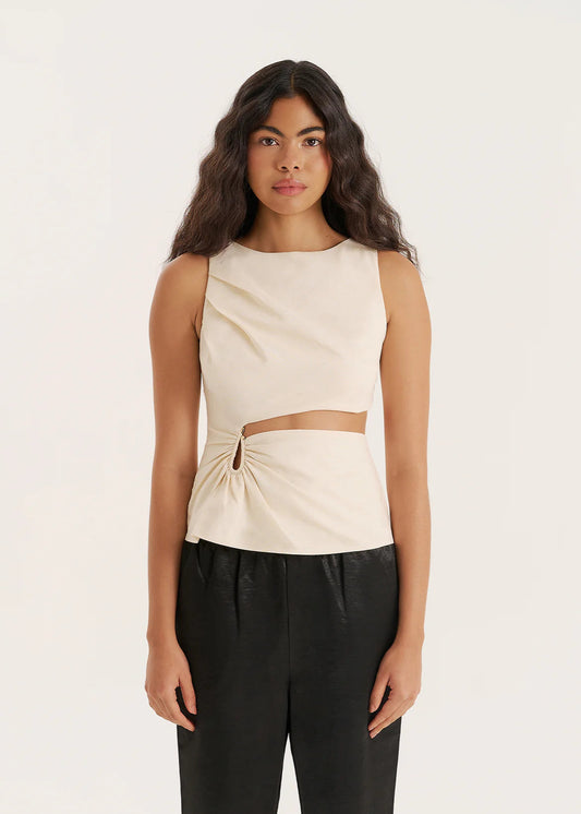 Woman wearing a beige sleeveless top with a cutout design and black pants on a white background