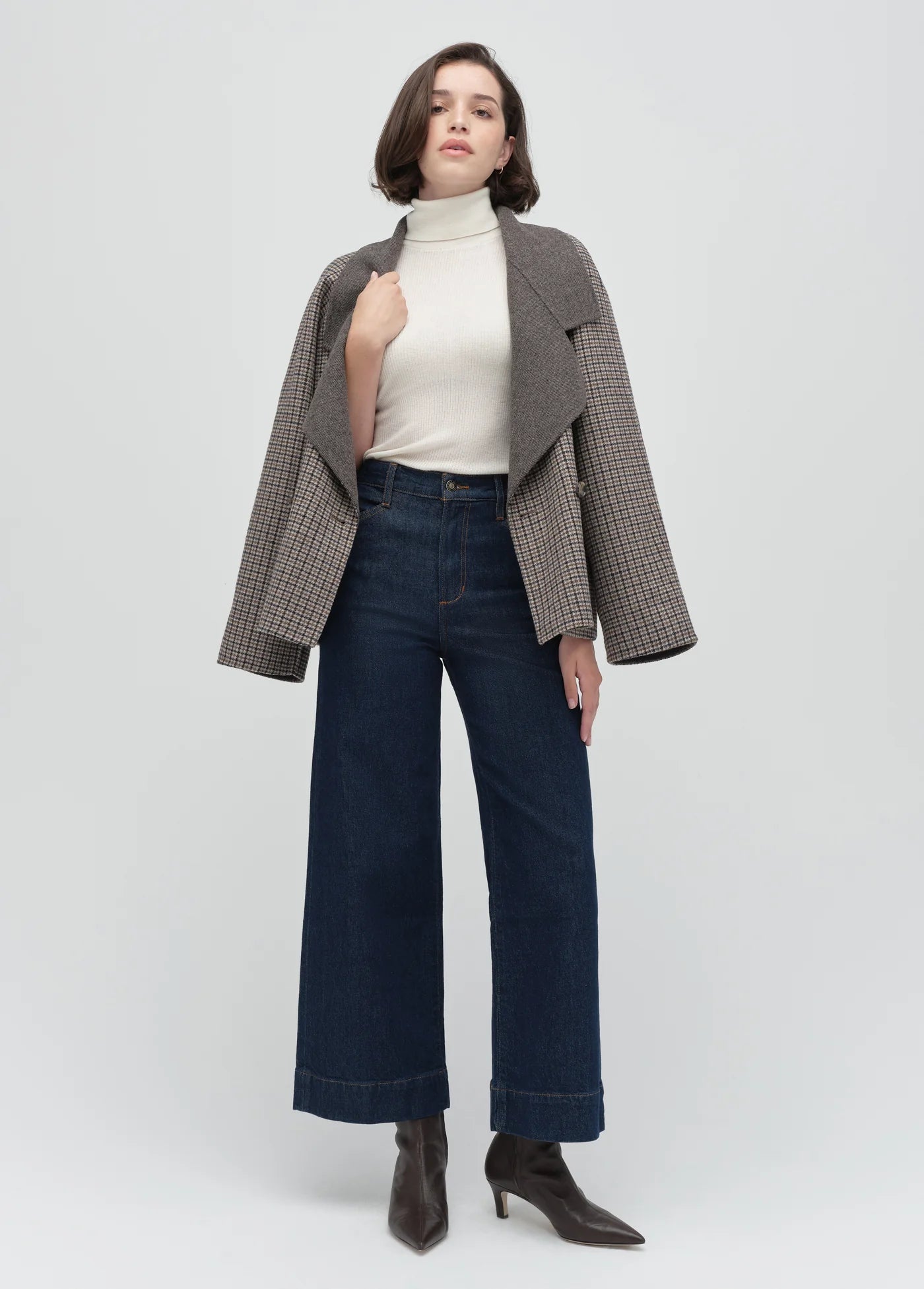 Woman wearing a plaid blazer, white turtleneck, dark jeans, and black boots on a white background